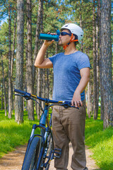 Cycling Fitness: An adult sport enthusiast in sportswear paused riding to drink water, amidst an urban park in surrounding by pine trees. This photo is part of a series themed by outdoor activities.