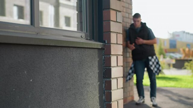 blurred view of young male student in casual outfit leaning on brick building corner while inhaling from vape device and coughing on sunny day with urban street and building background