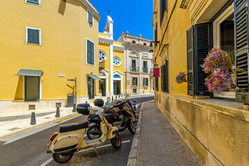 Narrow street with scooters parked against a backdrop of vibrant yellow buildings and historic architecture in Mahon, Spain.