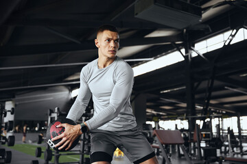Horizontal photo of fitness man model exercising with medicine ball at gym. Young strong guy doing workout using fitness ball. Strength training, discipline, and fitness lifestyle concept