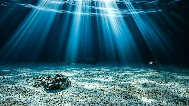 Sun rays pierce deep blue water, illuminating a sandy ocean floor with scattered rocks in a serene underwater vista