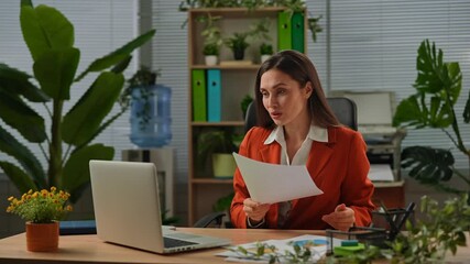 Older adult businesswoman works at green office with plants, woman sitting at desk working on laptop, talking online by video call, reading documents. - Powered by Adobe