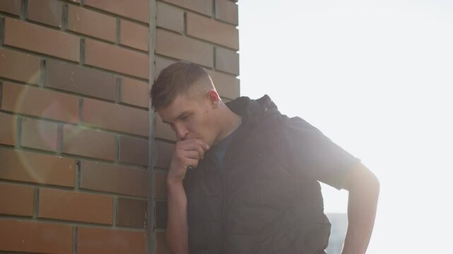 white man leaning on brick wall under sun flare coughing into hand after inhaling vape device emitting thick smoke wearing black puffer vest by urban apartment backdrop lens flare effect for moody