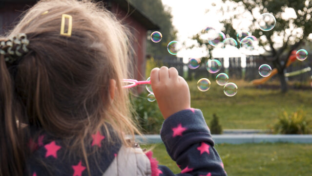 Young girl joyfully blowing soap bubbles in a backyard creates a playful atmosphere. Dressed in pink and blue jacket, she enjoys sunny day surrounded by green grass at yard on children playground