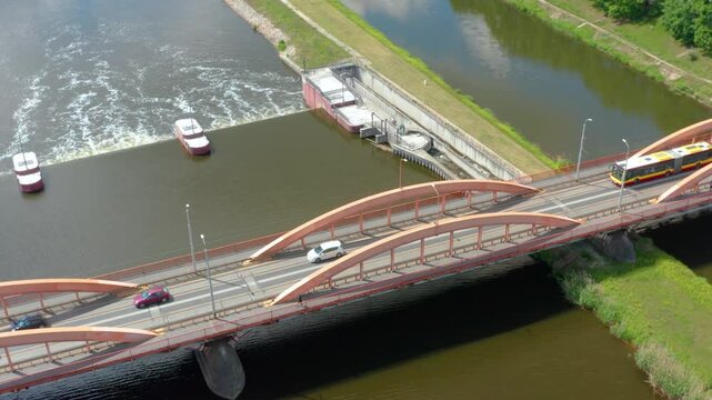 Tracking dDrone footage showing a red-arched bridge in Wroclaw, Poland, spanning over the Oder River with vehicles and a public bus in motion, highlighting urban infrastructure and transportation.