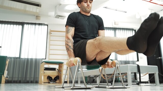 Man practicing L-sit exercise on pilates reformer to build core strength and upper body control at gym. Sporty man doing knee tuck on the reformer for strengthening core, arm, shoulder. Habituate.