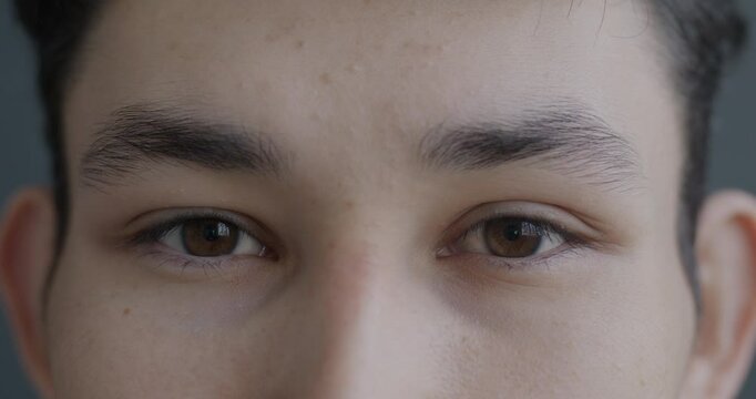 Close-up slow motion portrait of young Asian man's eyes looking at camera with no emotion on gray color background. People and face expression concept.