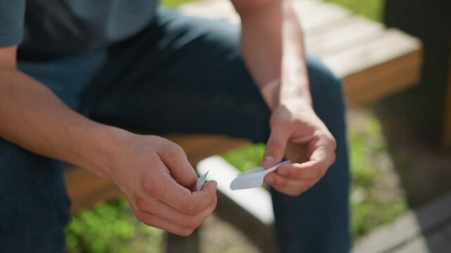 partial view of person seated placing nicotine patch on left arm while sitting on bench outdoors with soft greenery swaying gently in background