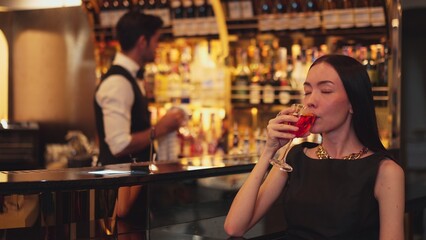 Beautiful guest looking at camera drinking at luxurious counter bar in mixed vodka Cosmopolitan red cocktail decorated with flower on bartender background in perfect atmosphere at nightclub. Vinosity.