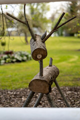 A creative wooden sculpture resembling a reindeer, crafted from tree logs and branches. The sculpture is placed in a natural outdoor setting, with a lush green lawn and colorful flowers in the backgro