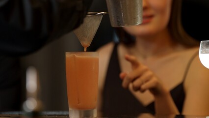 Beautiful woman guest looking at Paloma cocktail for preparation by bartender using strainer to...