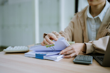 Fototapeta premium Hands of businessman hold pen for work in stacks of paper files, information search for business reports and heaps of incomplete documents is achieved on a laptop computer table in a modern office