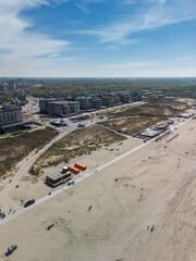 Aerial view of a modern beachside development with apartment buildings, sand dunes, and a wide sandy beach under a clear blue sky. People walk along the path and tracks mark the sand.