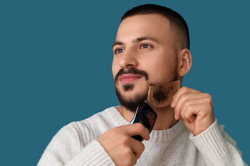 Handsome young bearded man with electric shaver and comb on blue background