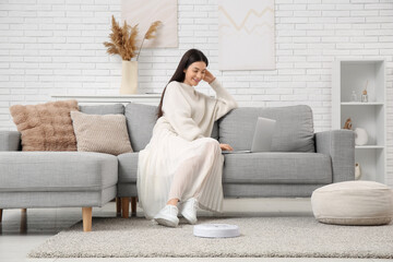 Young woman using laptop on sofa with robot vacuum cleaner at home
