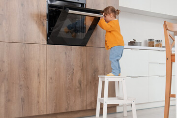 Cute little girl opening oven on stool in kitchen. Child at risk