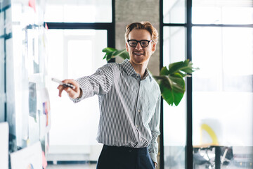 Smiling young male entrepreneur enthusiastically pointing at glass board with strategy charts...