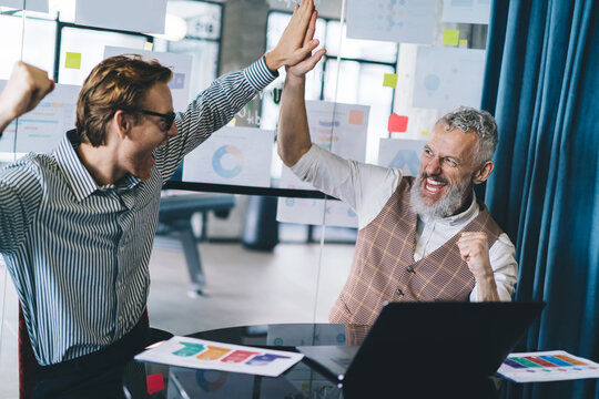 Joyful business success celebration with senior and young entrepreneurs giving high-five at desk, excited after achieving team goal in creative workplace