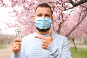 Sick young man in medical mask pointing at pills from allergy outdoors