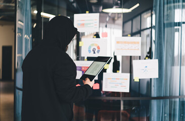 Anonymous hacker in black hoodie using tablet while analyzing corporate data charts on office glass...