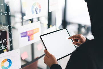 Suspicious man in hoodie interacts with blank tablet screen near analytics board, possibly masking identity during digital intrusion or unauthorized access attempt