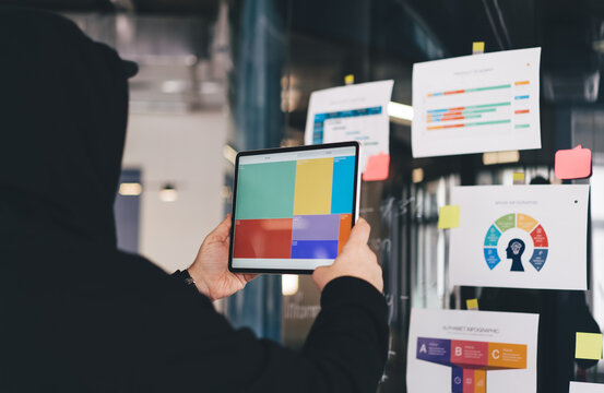 Cyber hacker analyzing colorful data blocks on tablet in front of strategic infographics, planning coordinated digital assault from within modern corporate environment