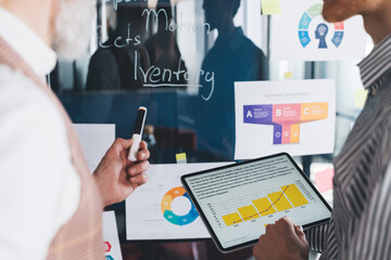 Close-up of businessmen analyzing data on digital tablet while discussing charts on glass wall, focused on numbers, progress, and effective business performance review