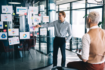 Young male professional presenting strategy on infographic wall while senior colleague listens...