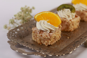 three delicate bite-sized square cake pieces topped with whipped cream and garnished respectively with orange slice, kiwi segment, and another citrus wedge, all artfully arranged on an ornate silver