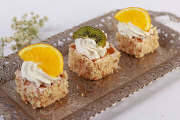 three delicate bite-sized square cake pieces topped with whipped cream and garnished respectively with orange slice, kiwi segment, and another citrus wedge, all artfully arranged on an ornate silver