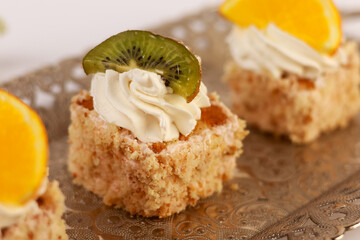 three delicate bite-sized square cake pieces topped with whipped cream and garnished respectively with orange slice, kiwi segment, and another citrus wedge, all artfully arranged on an ornate silver