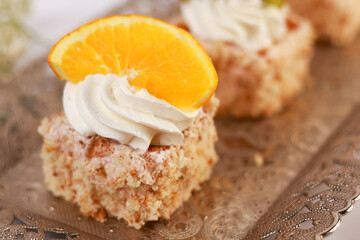 three delicate bite-sized square cake pieces topped with whipped cream and garnished respectively with orange slice, kiwi segment, and another citrus wedge, all artfully arranged on an ornate silver