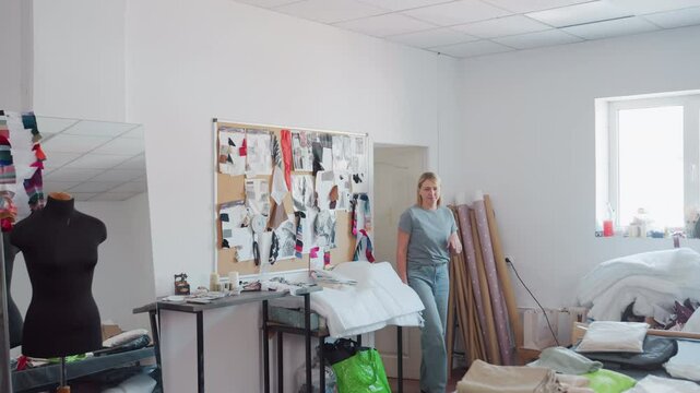 Designers workspace filled with colorful fabrics, sewing tools, design board, window light as two women walk in discussing ideas, scattered clothing designs from cluttered work surface