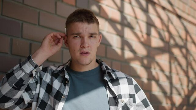 young man in black white check shirt returns cigarette to ear while gazing into distance from metal staircase landing against brick wall backdrop with dappled sunlight and lush greenery beyond