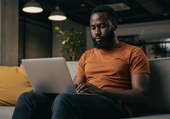 A black man is using a laptop while sitting on a gray sofa, working remotely in his home.
