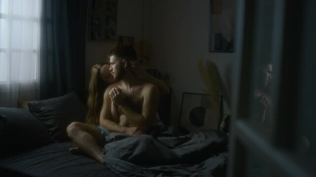 Full through-doorway shot of young Caucasian couple in underwear sitting in bed in morning and cuddling, woman embracing spouse from behind and stroking neck, they touching hands, deep feelings