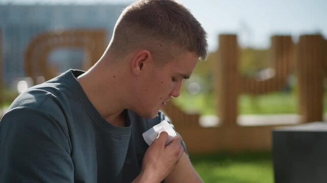 side view of young boy gently placing nicotine patch on upper arm while seated outdoors under bright sunlight with soft blur background