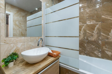 The bathroom is decorated with beige and brown tiles. A bathtub with a shower behind a glass...