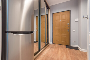 An entrance hall with a brown door in a small apartment. A wardrobe with mirrored doors and a two-chamber refrigerator.