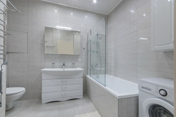 Modern bathroom interior design. Light tiles, white furniture. A bathtub with a shower behind a glass partition.