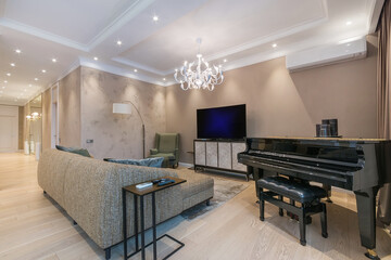 Exquisite interior of the living room in pastel colors. A soft gray sofa in front of the TV, a black piano. A luxurious chandelier.