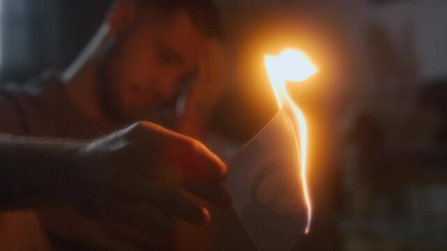 Close-up shot of young crying man burning photo of beloved woman at home, feeling depressed and vulnerable after painful breakup