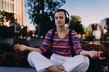 Calm young woman meditating with closed eyes and headphones on, sitting cross-legged in city park,...