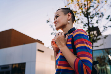 Fototapeta premium Young woman gently holds her headphones while smiling softly in warm afternoon light, enjoying a quiet city moment in vibrant striped clothes and minimalist style