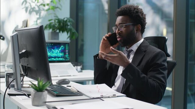 Confused African American man employee talking mobile phone businessman business cellphone looking computer monitor pc worker manager male call problem trouble puzzled guy consulting client in office
