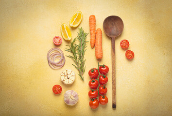 Cherry tomatoes, garlic, lemon slices, carrots, red onions, and rosemary sprig arranged alongside a wooden spoon on a yellow backdrop, culinary creativity and healthy eating concept