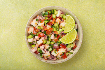 Fresh and vibrant peruvian seafood ceviche with shrimps, octopus, tomatoes, cucumber and parsley, served with lime in a bowl on a light green background from above, dish of Peru