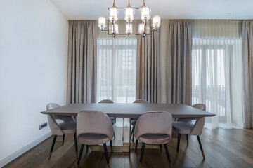 The interior of the dining room in a luxury apartment with panoramic windows. Beautiful chandelier over the dining table with gray chairs.