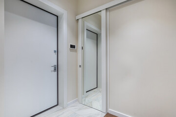 A bright corridor with a white entrance door. The intercom screen is on the wall. A small mirrored sliding door.