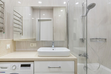 The interior of a small bathroom. Mirrored cabinet over the sink, built-in washing machine. A bathtub with a shower behind a glass partition.